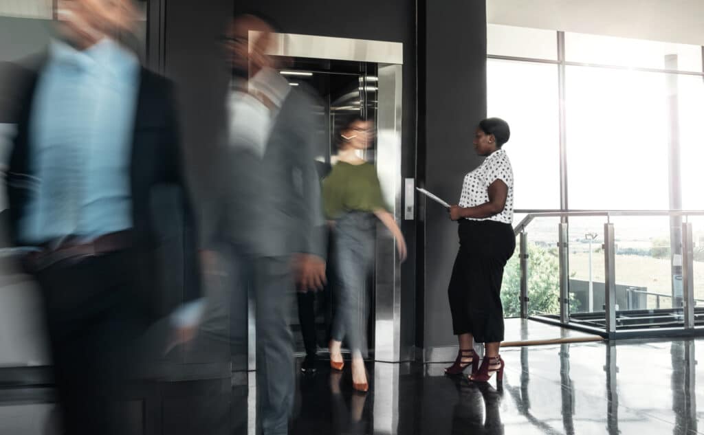 Business woman standing with paperwork, waiting to enter a busy elevator lift. With people walking out in a blur, showing motion.
