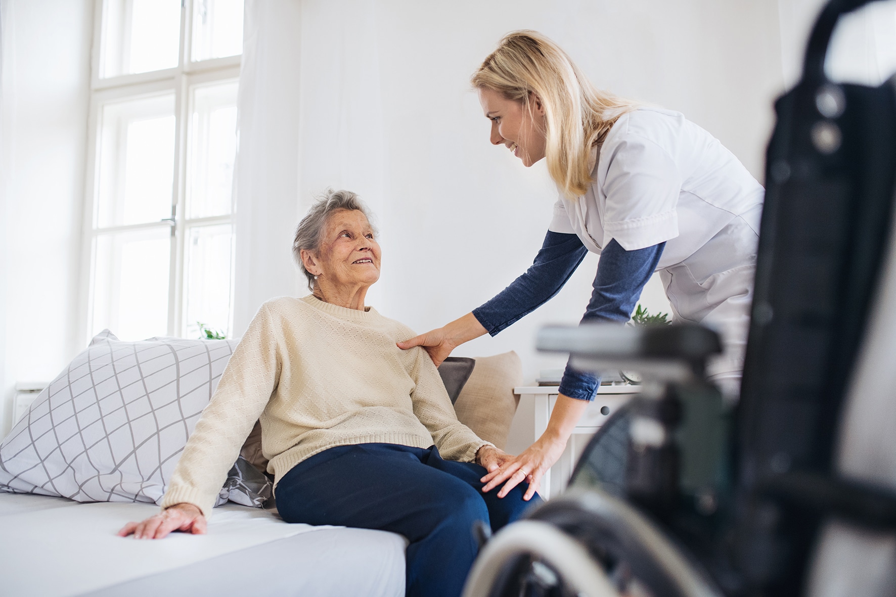 A health visitor talking to a senior woman sitting on bed at home.