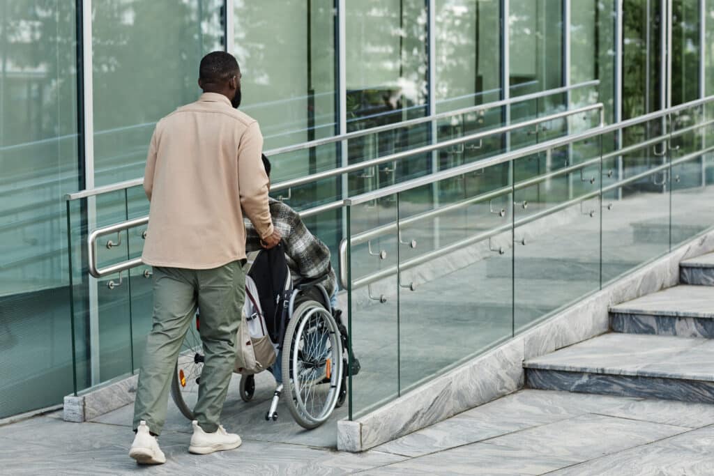 Back view of adult man assisting partner in wheelchair going up accessibility ramp in city, copy space