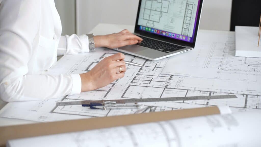 Architect studying detailed building plans at desk, using compass, ruler and laptop for technical drafting and digital design to develop modern residential and urban construction projects