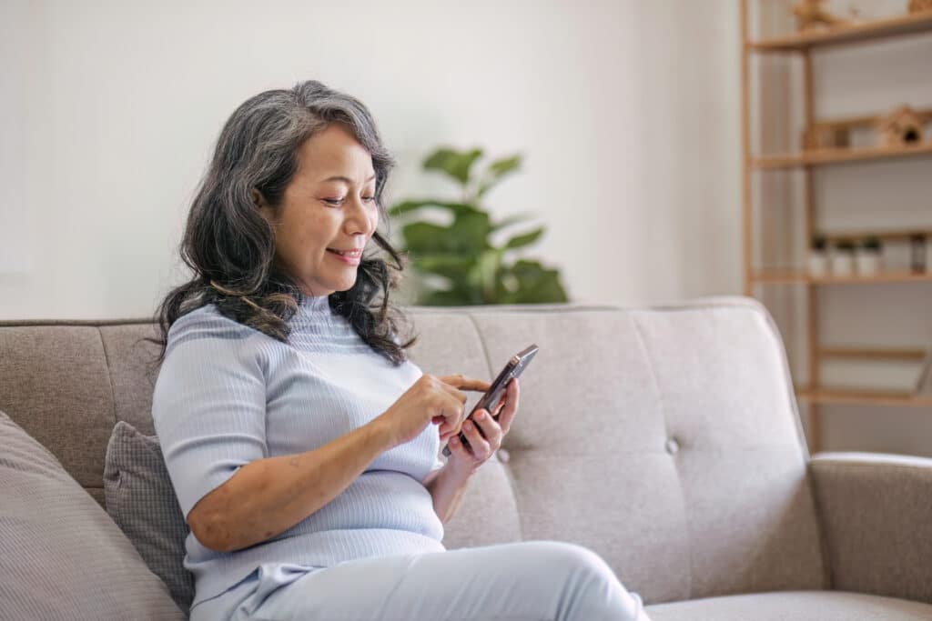 Portrait, Happy 60s retired Asian woman using her phone on a sofa in the living room. chatting, scrolling on the phone.